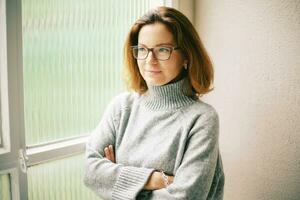 intérieur portrait de magnifique 40 année vieux femme relaxant suivant à fenêtre, portant chaud gris arrêtez-vous et des lunettes photo