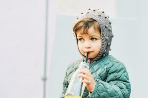 Extérieur portrait de mignonne peu fille en buvant jus de bouteille, portant chaud veste photo