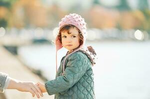 Extérieur portrait de mignonne bambin fille en marchant par le lac, en portant celui de maman main, à la recherche retour plus de le épaule, portant pièce veste et rose chapeau, du froid temps photo