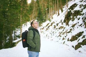 Extérieur portrait de milieu âge 55 - 60 année vieux homme randonnée dans hiver forêt, portant chaud veste et noir sac à dos photo