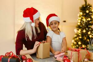 vacances, fête, famille et gens concept - content mère et peu fille dans Père Noël assistant Chapeaux avec cadeau boîte plus de vivant pièce et Noël arbre Contexte photo