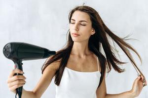 portrait d'une magnifique fille brune soufflant du vent sur ses cheveux mouillés tout en tenant un sèche-cheveux dans les mains et fermant les yeux avec plaisir. stock photo