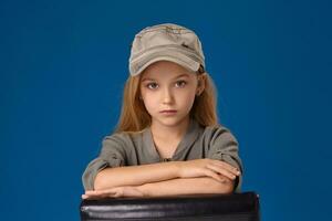 peu fille dans une casquette avec gris yeux et blond cheveux séance sur une chaise photo