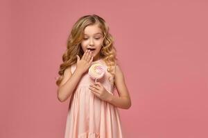 peu fille avec une blond frisé cheveux, dans une rose robe est posant avec une bonbons photo