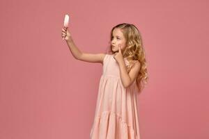 peu fille avec une blond frisé cheveux, dans une rose robe est posant avec une bonbons photo