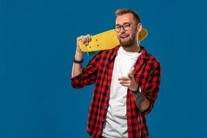 charismatique de bonne humeur Jeune barbu homme habillé dans à carreaux chemise, blanc T-shirt et lunettes, avec Jaune planche à roulette dans le sien mains. studio coup avec bleu Contexte photo