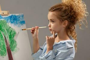 charmant école fille est La peinture avec une aquarelle brosse sur un chevalet, permanent sur une gris Contexte. photo