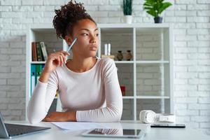 Jeune femme mulâtre sérieuse est assise et regarde pensivement sur le côté en touchant avec un stylo ses joues photo