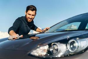 vue portrait à la taille du jeune propriétaire masculin heureux nettoyant sa voiture noire avec un chiffon tout en se tenant dans la rue pendant la journée ensoleillée. stock photo