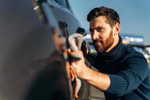 homme essuyant sa voiture dans la rue. lavage de détail de voiture pendant la journée ensoleillée. bel homme barbu dans des vêtements décontractés laver les portes et le capot de la voiture avec un chiffon en microfibre photo