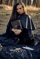 sorcière dans noir robe avec cap et capot. posant dans pin forêt. séance sur foncé couverture avec livres, bougies et runes sur il. plein longueur. photo