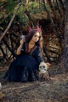 sorcière dans noir, longue robe, avec rouge couronne dans sa longue cheveux. posant avec balai et crâne dans pin forêt. sorts, la magie et la sorcellerie. plein longueur. photo