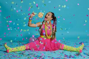 charmant fille avec une multicolore tresses coiffure et brillant se maquiller, posant dans studio avec sucette, air des ballons et confettis contre une bleu Contexte. photo