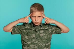 fermer portrait de une blond adolescent garçon dans une vert chemise avec paume impression posant contre une bleu studio Contexte. concept de sincère émotions. photo