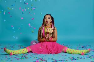 charmant fille avec une multicolore tresses coiffure et brillant se maquiller, posant dans studio avec sucette, air des ballons et confettis contre une bleu Contexte. photo