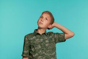 fermer portrait de une blond adolescent garçon dans une vert chemise avec paume impression posant contre une bleu studio Contexte. concept de sincère émotions. photo