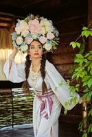 brunette fille dans une blanc ukrainien authentique nationale costume et une couronne de fleurs est posant contre une vert cour. fermer. photo