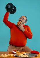 fermer portrait de une âge moyen homme avec barbe, habillé dans une rouge col roulé, posant avec des hamburgers et français frites. bleu Contexte. vite aliments. photo