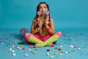 charmant fille avec une multicolore tresses coiffure et brillant se maquiller, posant dans studio avec sucette, air des ballons et confettis contre une bleu Contexte. photo