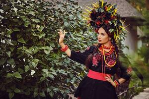 brunette fille dans noir et rouge brodé ukrainien authentique nationale costume et couronne de fleurs est en marchant seul et posant dans une vert jardin. photo