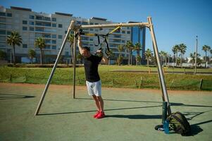 Jeune adulte athlétique gars formation bras en utilisant suspension aptitude les bretelles pendant un Extérieur faire des exercices dans Urbain des sports sol photo