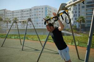 travail dur. côté vue Jeune athlétique homme avec parfait corps dans tenue de sport Faire pousser UPS avec aptitude les bretelles en plein air photo