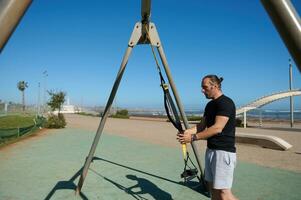 actif Jeune sportif pendaison suspension les bretelles sur une barre transversale, prêt pour une poids faire des exercices dans Extérieur des sports sol photo