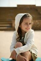 fermer élégant peu enfant fille séance sur sa bleu planche à roulette, souriant rêveusement à la recherche loin, habillé dans beige sweat à capuche photo