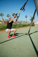 Jeune adulte sportif dans noir T-shirt et gris short exercice avec suspension les bretelles dans le Urbain des sports sol photo