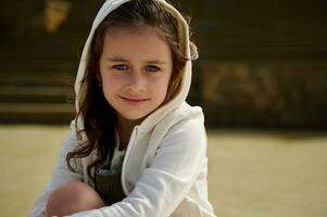 fermer peu enfant fille avec capuche sur sa diriger, souriant à la recherche en toute confiance à caméra, séance sur une planche à roulette Extérieur photo