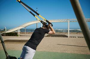 arrière vue de Jeune adulte athlétique homme en utilisant suspension les bretelles tandis que exercice force dans le en plein air terrain de sport photo