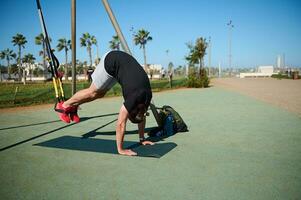 actif Jeune adulte athlétique homme Faire craquements, permanent sur le sien mains tandis que exercice avec suspension les bretelles photo