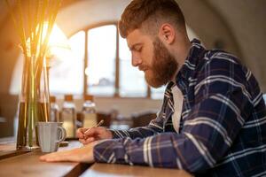 Jeune barbu homme d'affaire est assis dans café, Accueil à table et écrit dans carnet. homme est fonctionnement, en étudiant. photo