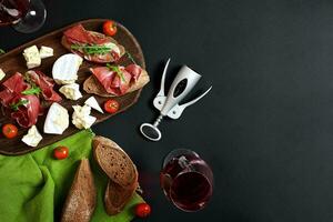 délicieux apéritif à du vin - jambon, fromage, baguette tranches, tomates, servi sur une en bois conseil, et verre avec rouge du vin sur noir surface photo