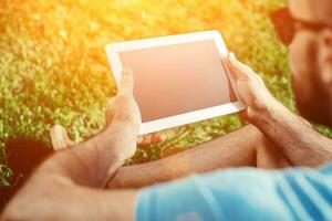 fermer de décontractée habillé Jeune homme en utilisant moderne numérique tablette tandis que séance à le parc, Soleil lumière photo