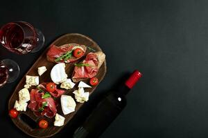 délicieux apéritif à du vin - jambon, fromage, baguette tranches, tomates, servi sur une en bois conseil, et verre avec rouge du vin sur noir surface photo