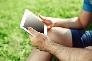 fermer de décontractée habillé Jeune homme en utilisant moderne numérique tablette tandis que séance à le parc, Soleil lumière photo