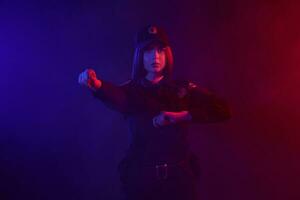 rousse femelle police officier est posant pour le caméra contre une noir Contexte avec rouge et bleu rétro-éclairage. photo