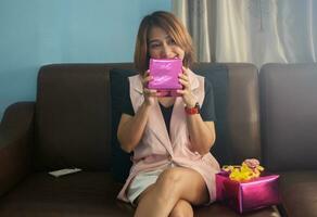 femme séance sur canapé en portant cadeau boîte dans de face de visage et souriant bonheur à maison, gens mode de vie concepts photo
