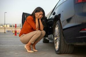 portrait de une désespoir femme accroupi suivant à sa voiture avec plat pneu, problèmes sur le route, dégonflé pneu. photo