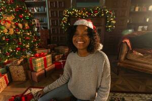 joyeux Noël. africain américain femme souriant près Noël arbre dans classique foncé intérieur. content fille dans Père Noël chapeau dans vivant pièce avec cheminée Noël arbre cadeau des boites. Noël veille à maison. photo