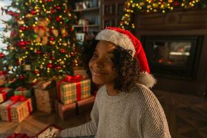 joyeux Noël. africain américain femme souriant près Noël arbre dans classique foncé intérieur. content fille dans Père Noël chapeau dans vivant pièce avec cheminée Noël arbre cadeau des boites. Noël veille à maison. photo