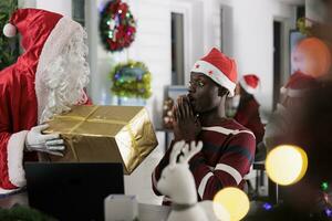 africain américain employé surpris à voir Noël cadeau offert par collaborateur costumé comme Père Noël claus pendant hiver vacances saison. ouvrier offre Noël présent à collègue dans de fête orner Bureau photo