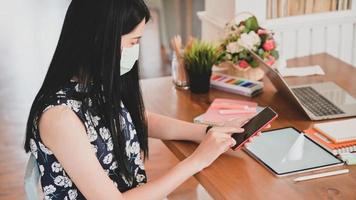 une femme portant un masque médical utilise un smartphone avec une tablette, un ordinateur portable et du matériel de bureau sur le bureau.