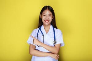 portrait de une magnifique Jeune femme dans une Jaune arrière-plan, amical magnifique femme portant une médecins uniforme et montrer du doigt tandis que portant une stéthoscope. photo