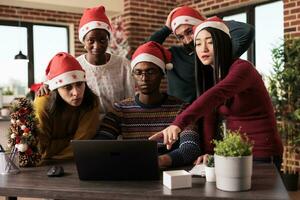 diverse collègues de travail portant Père Noël Chapeaux vérification annuel rapport sur portable ensemble dans décoré Bureau sur Noël veille. entreprise des employés équipe travail pour date limite pendant hiver vacances photo