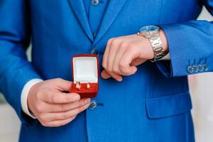 une homme détient mariage anneaux dans une rouge boîte dans le sien mains et regards à le sien regarder photo