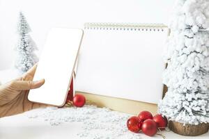 doux mains berceau une téléphone intelligent avec une blanc écran maquette et blanc calendrier avec une de fête scène de une magnifique Noël arbre orné, avec Noël des balles, pin cônes, et une rouge maison cadeau boîte. photo