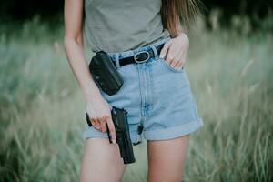 fille dans denim short et avec une pistolet dans le sien main posant dans le champ. proche en haut photo