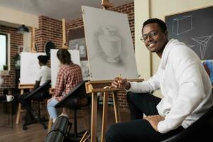 content africain américain gars séance à chevalet dans salle de cours profiter fabrication art pendant groupe dessin classe, Créatif espace pour social connexion. souriant homme apprentissage Comment à dessiner à art atelier photo